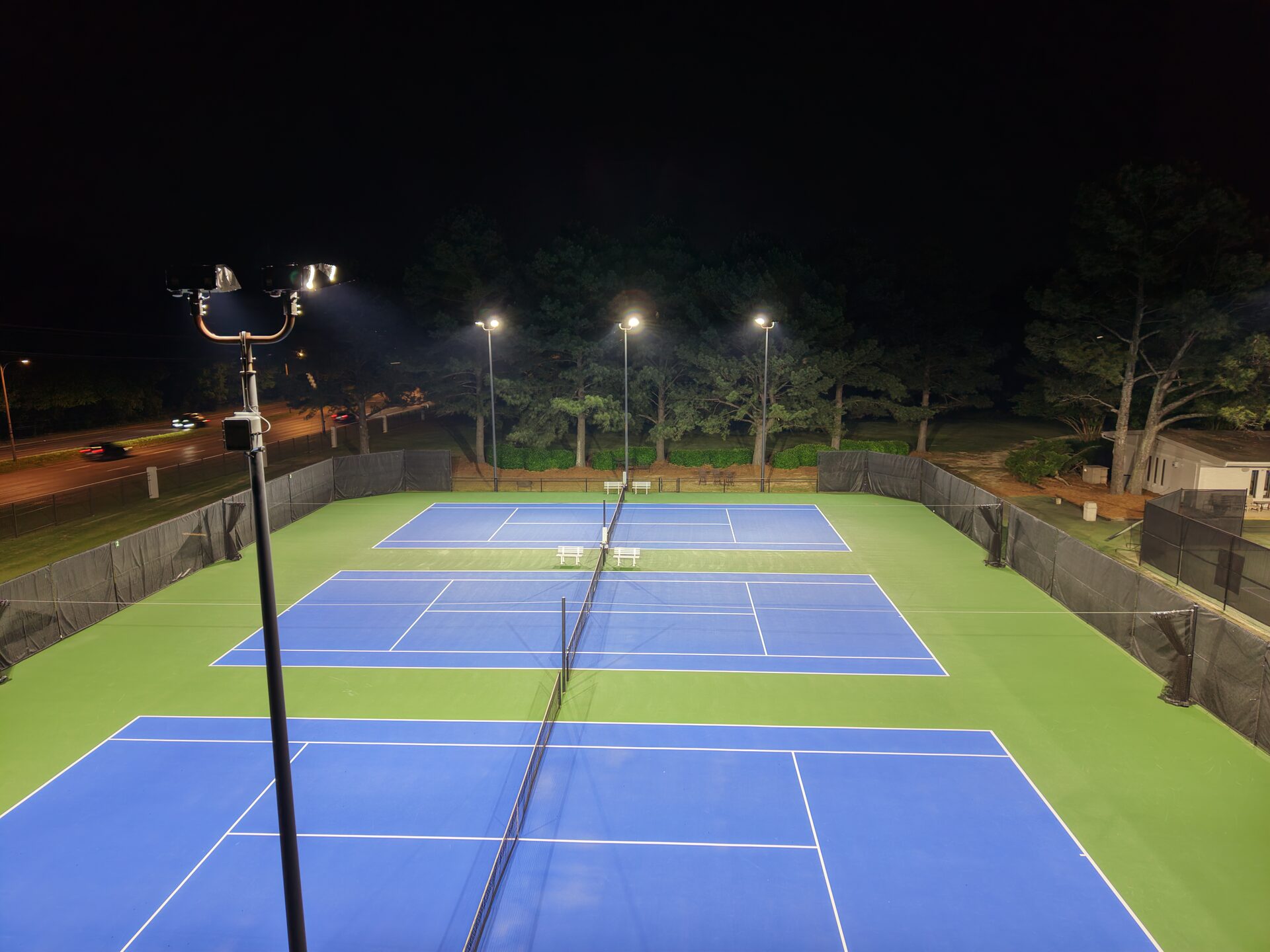 Ground-level player perspective view down the length of the tennis courts, with even illumination and no dark baseline areas